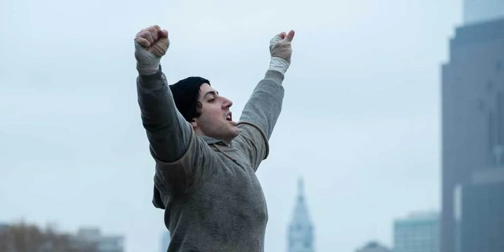 شاب يرتدي قبعة وقفازات يرفع ذراعيه بفخر في مشهد من فيلم "I Play Rocky" المعروض في مهرجان تورنتو السينمائي TIFF 2023.